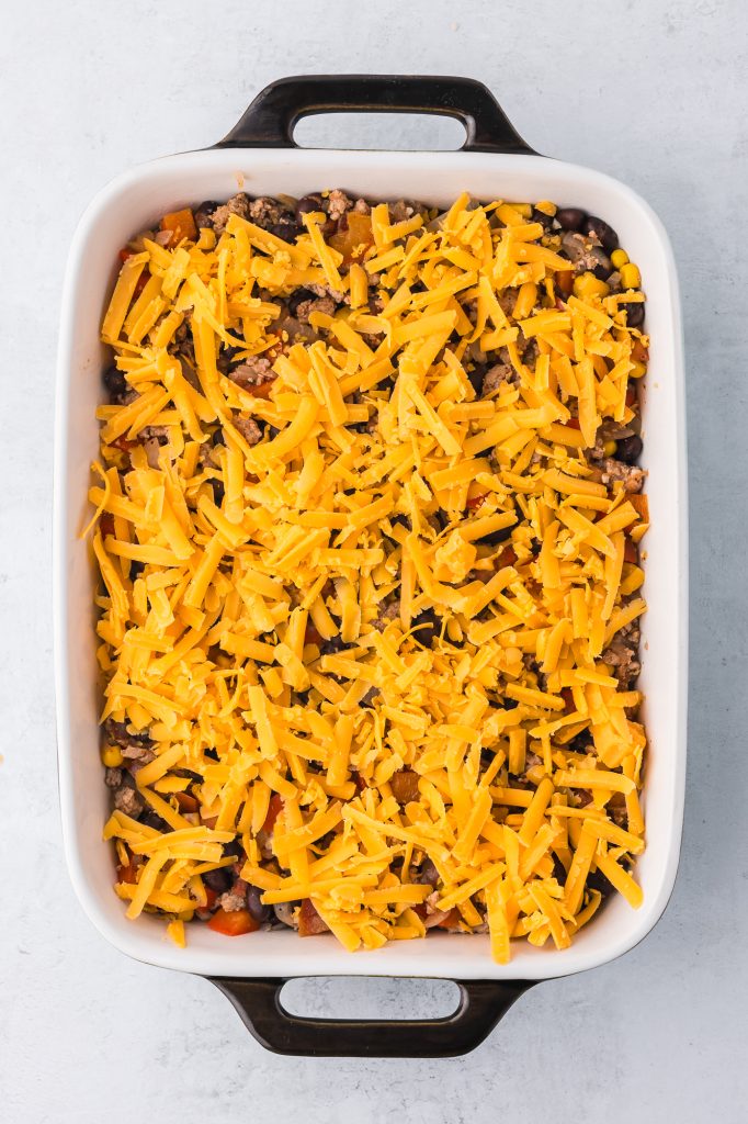 A casserole dish filled with cooked ground meat, peppers, onions, garlic, corn, and black beans covered with shredded cheese.