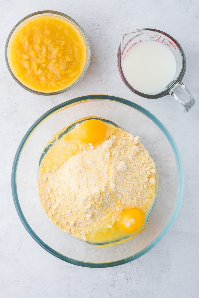 A glass bowl with Jiffy Mix and eggs, with smaller bowls of milk and creamed corn to the side.