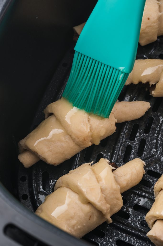 A raw Nutella Croissant crescents in an air fryer basket getting brushed with melted butter.