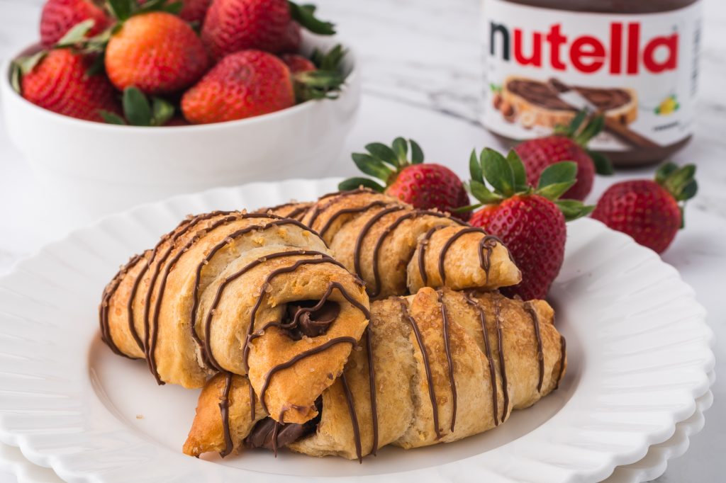 Three Nutella-drizzled Nutella Croissants on a white plate with strawberries in the background.