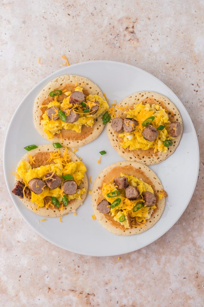 A plate of four pancakes with scrambled eggs, sliced sausage, shredded cheese, and sliced green onion on top.