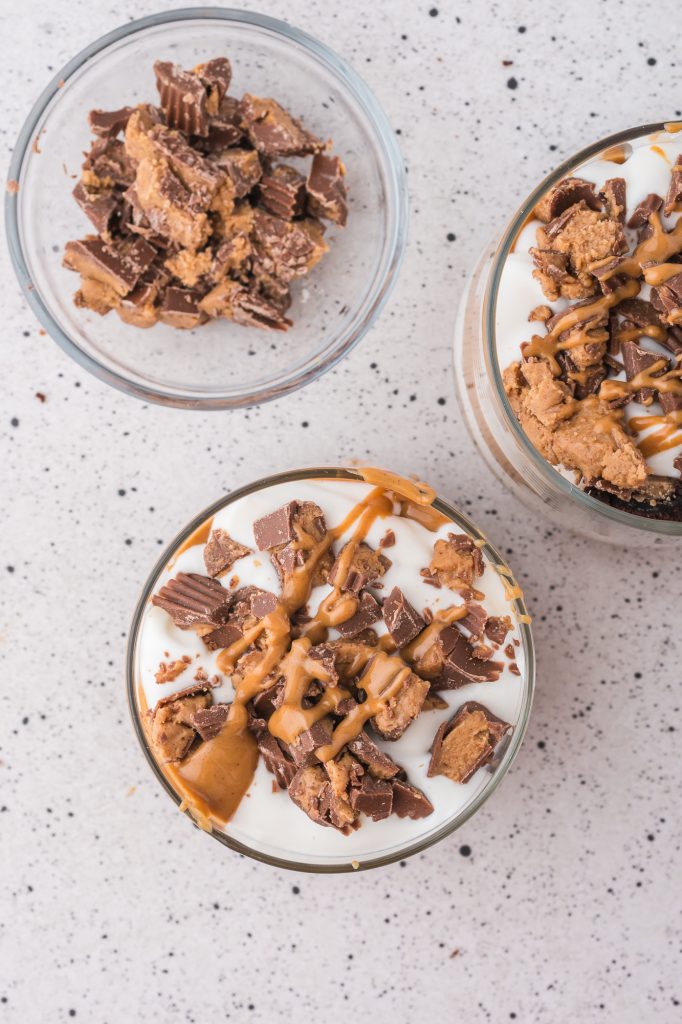 Two Peanut Butter Brownie Parfaits topped with chopped peanut butter cups, next to a bowl of chopped peanut butter cups.