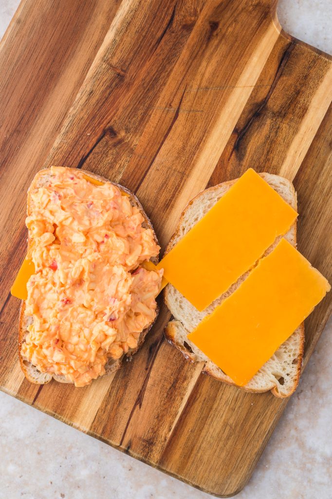 Two slices of bread with slices of sharp cheddar cheese on them, the slice on the left also has a heap of pimento cheese on it.