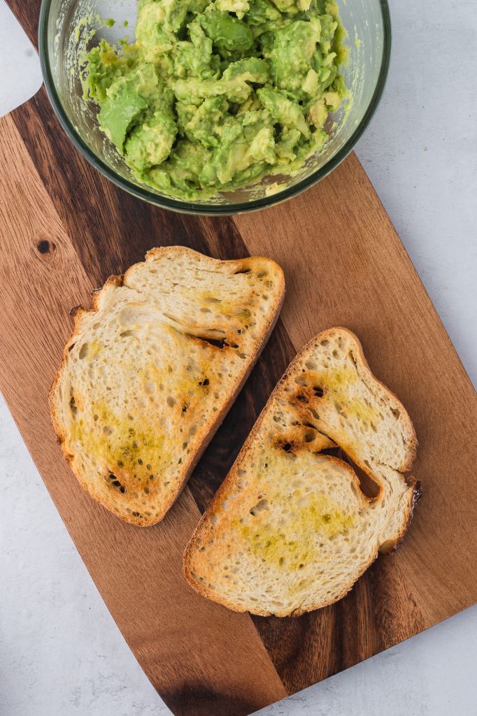 Two slices of toasted bread next to a bowl of mashed avocado.