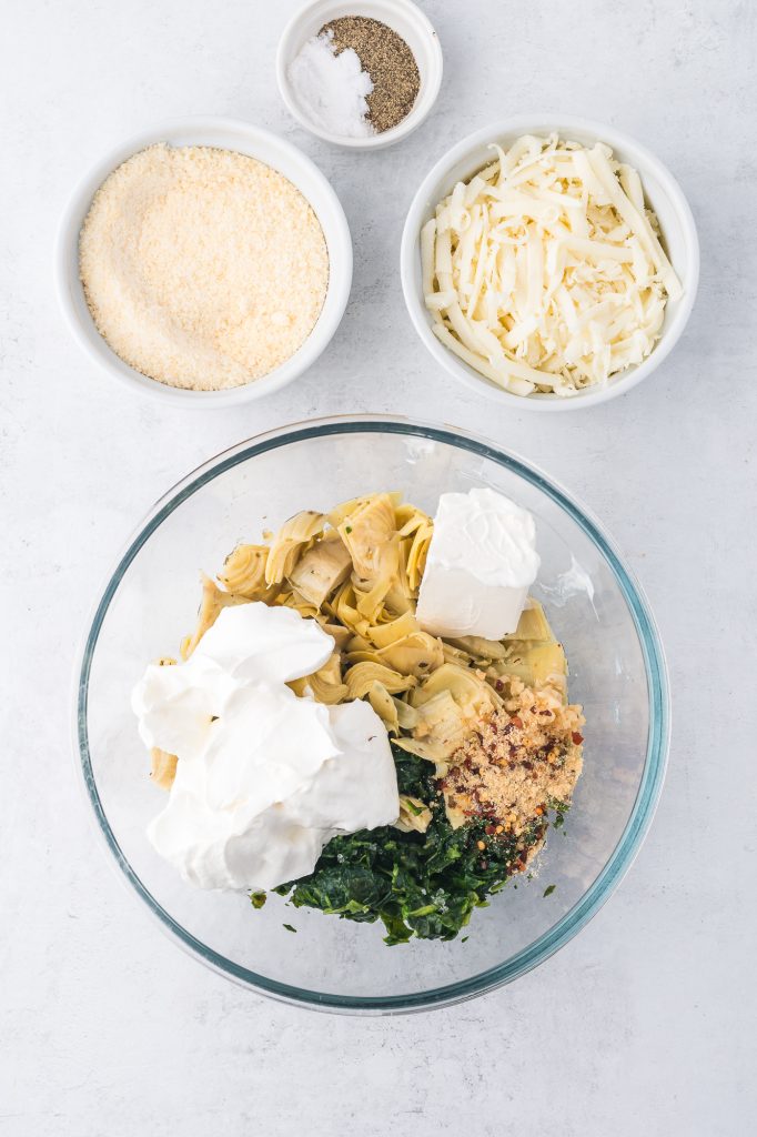 A glass bowl with the ingredients for No Mayo Spinach Artichoke Dip in it before mixing.