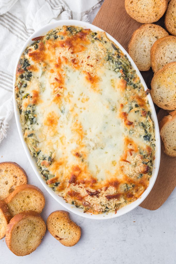 The baked No Mayo Spinach Artichoke Dip spread in a casserole dish surrounded by small crostinis.