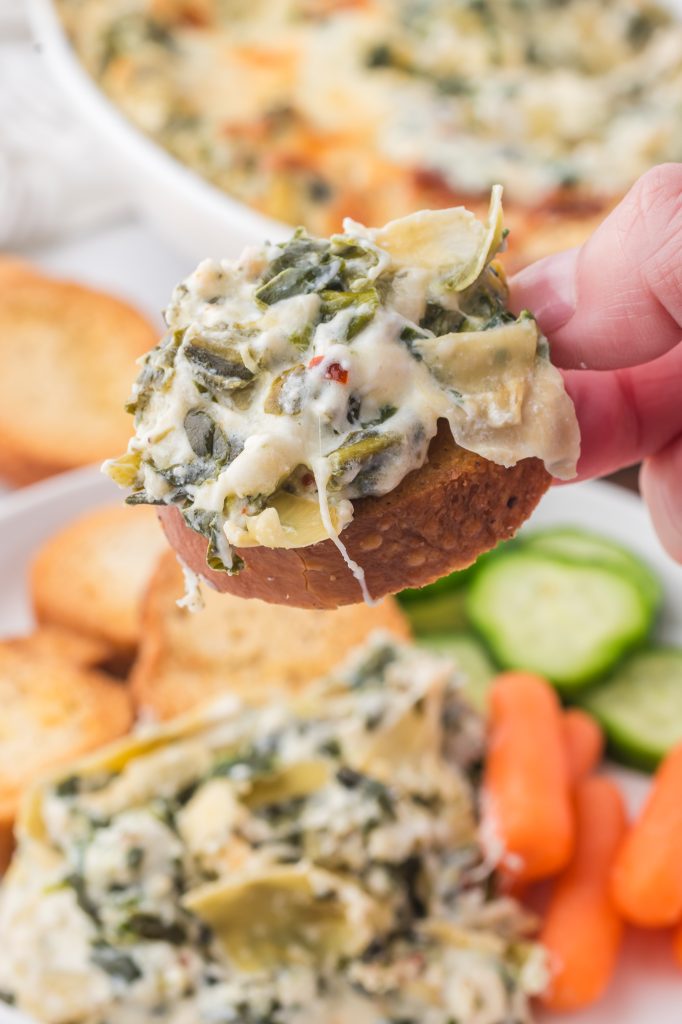 A close-up of a crostini being dipped into No Mayo Spinach Artichoke Dip.