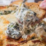 A close-up of a crostini being dipped into No Mayo Spinach Artichoke Dip.