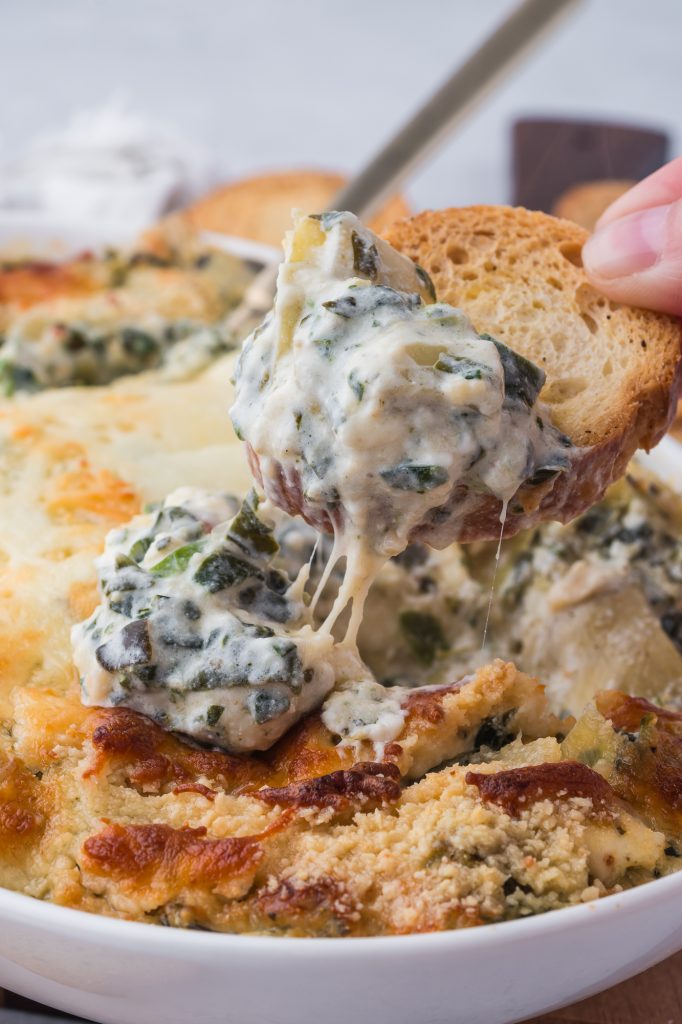 A close-up of a crostini being dipped into No Mayo Spinach Artichoke Dip.