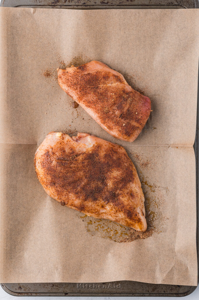 Two seasoned chicken breasts on a parchment lined baking sheet.