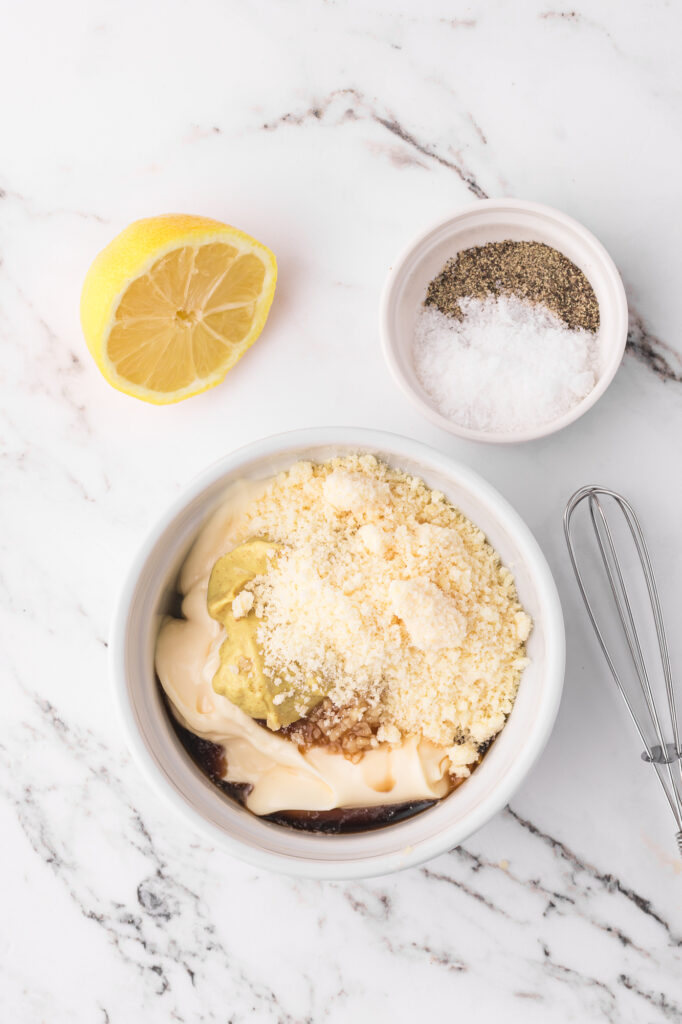 A small bowl filled with the ingredients for Homemade Caesar Dressing next to a smaller bowl of salt and pepper.