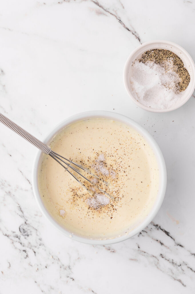 A small bowl filled with the blended ingredients for Homemade Caesar Dressing next to a smaller bowl of salt and pepper.