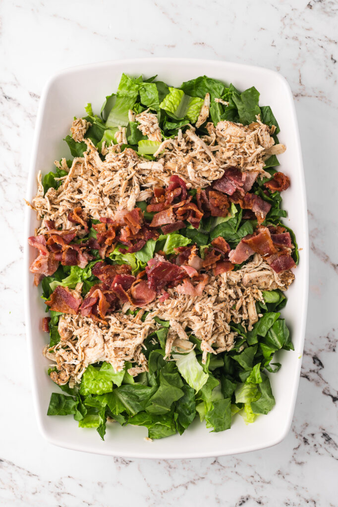 Some shredded chicken and bacon placed on a bed of chopped romaine in a large bowl.