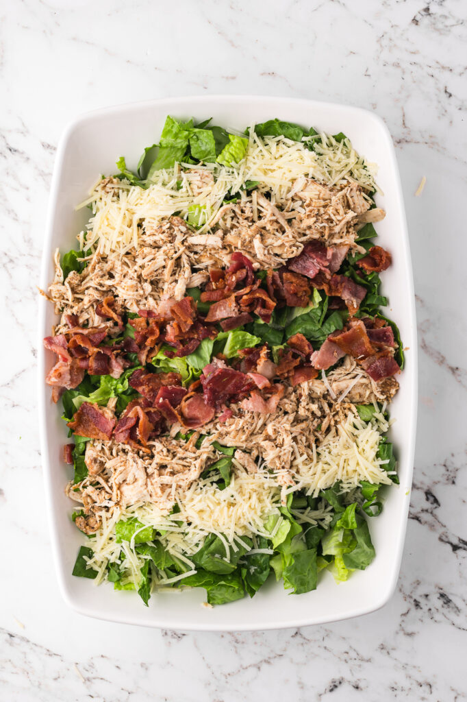 Some shredded chicken, bacon, and Parmesan cheese placed on a bed of chopped romaine in a large bowl.