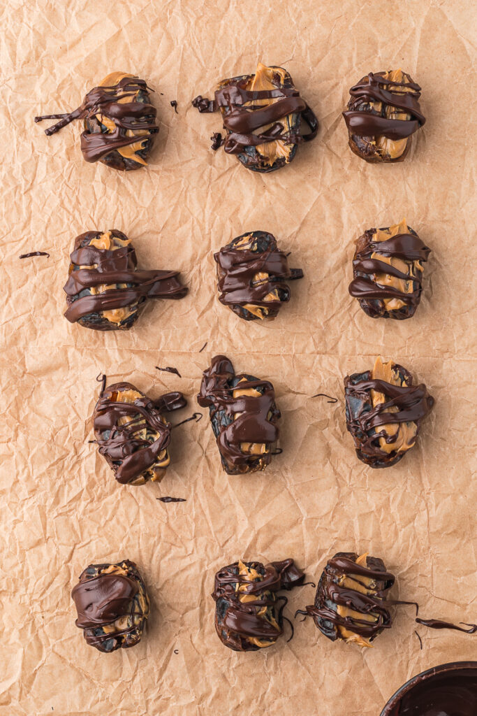 A piece of parchment paper holding 12 pitted dried dates stuffed with peanut butter and drizzed with chocolate.