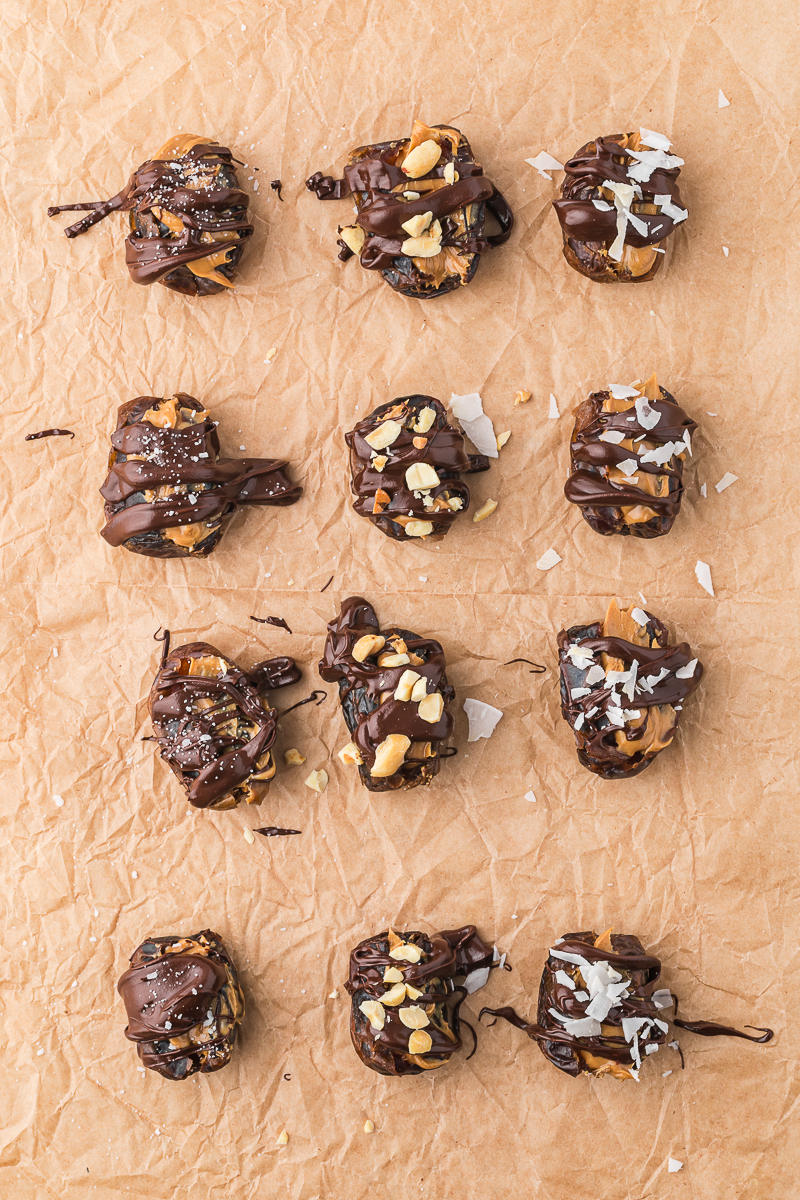 A piece of parchment paper holding 12 pitted dried dates stuffed with peanut butter, drizzed with chocolate, and topped with a variety of topping like coconut flakes and sea salt.