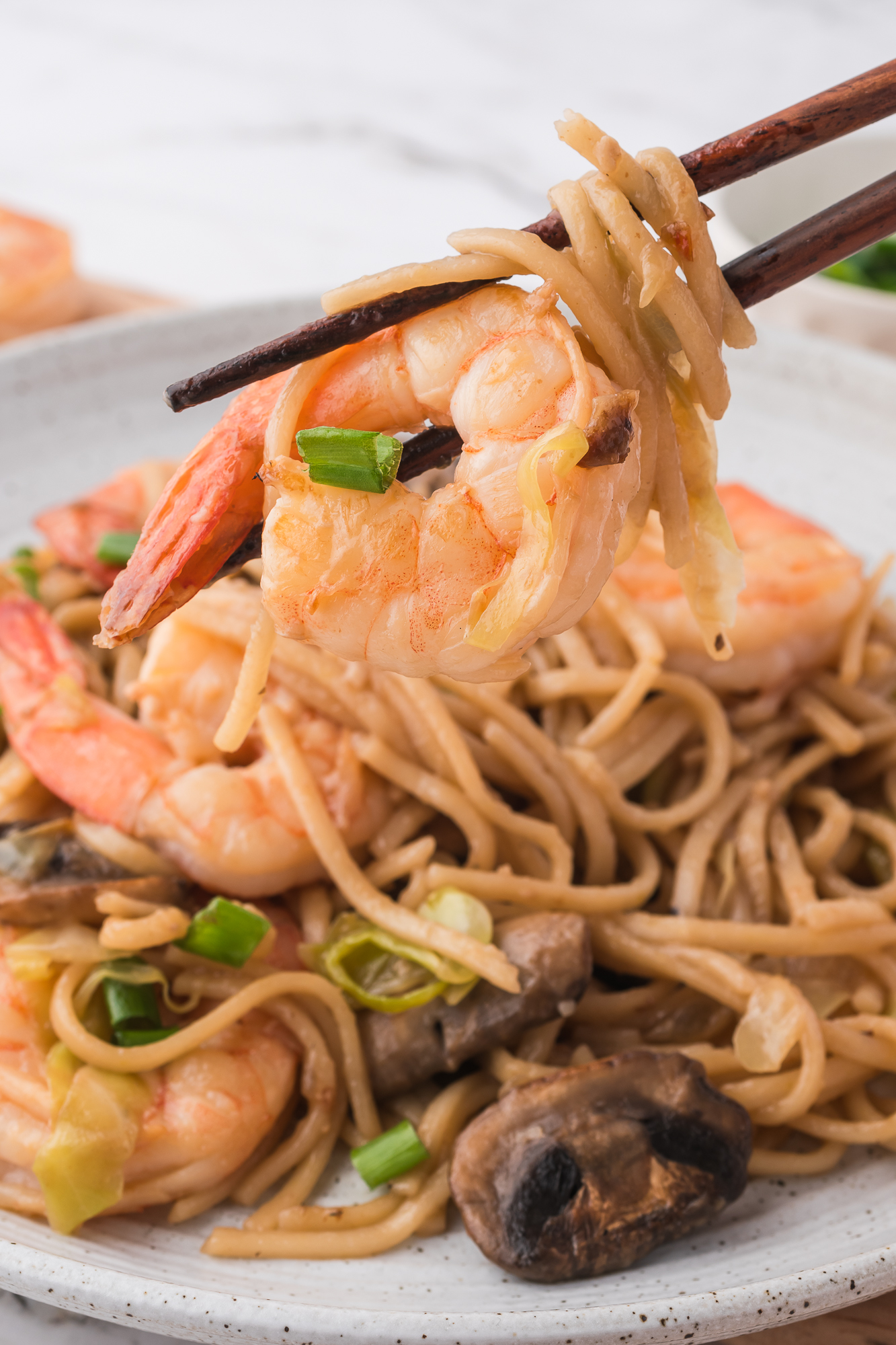 A close of of shrimp between chopsticks hovering over a plate of Shrimp Chow Mein.