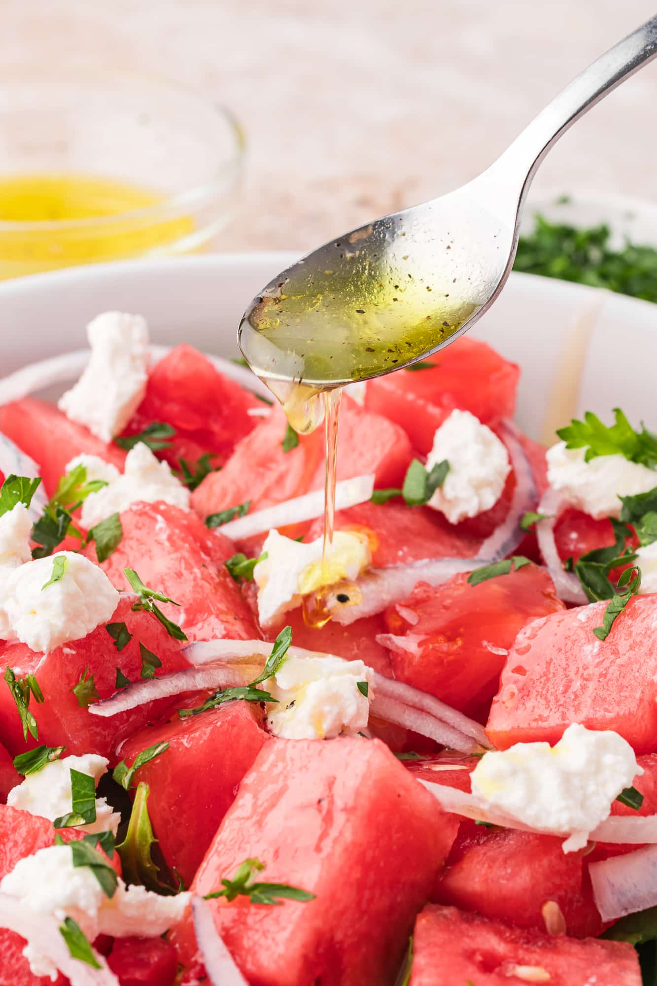 A close up of a spoon pouring dressing over a white bowl containing Watermelon Goat Cheese Salad.