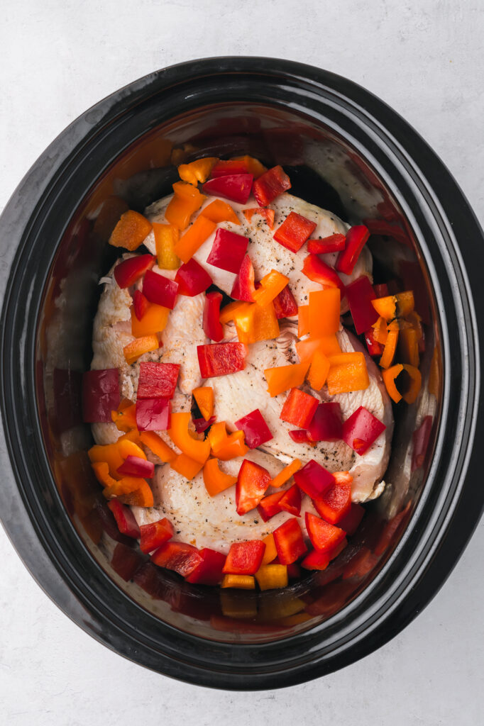 A slow cooker filled with seared chicken breast, topped with chopped bell peppers.