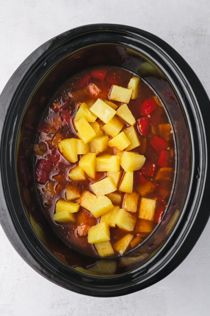 A slow cooker filled with seared chicken breast, topped with chopped bell peppers, sauce, and pineapples.