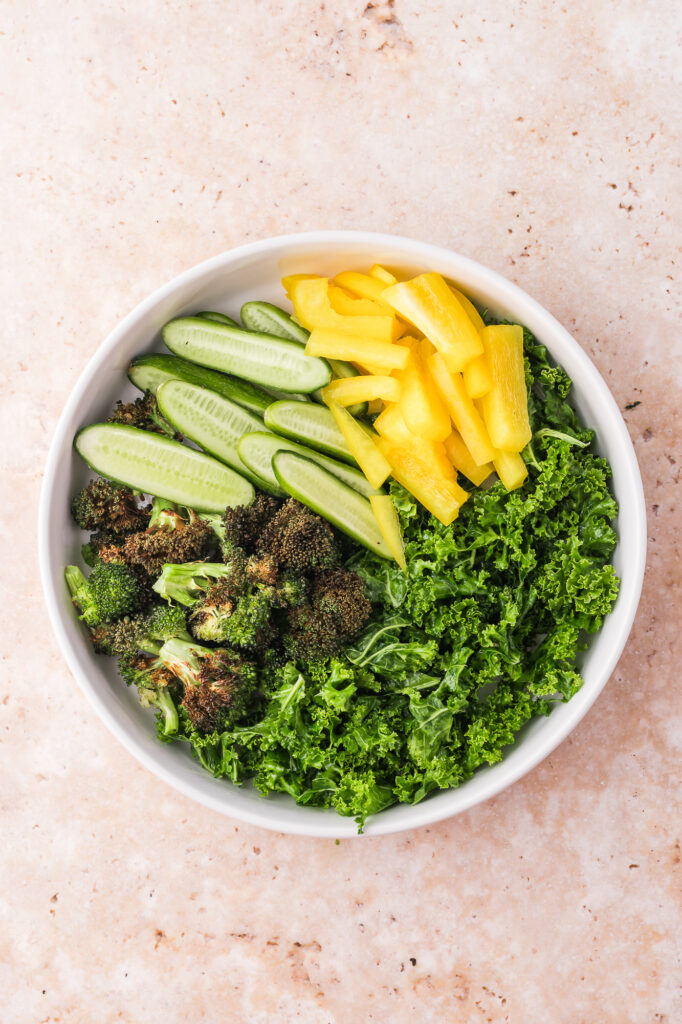 A white bowl of kale, roasted broccoli, cucumber, and yellow pepper.