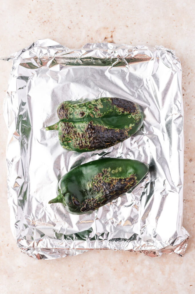 Two charred poblano peppers on a foil-lined sheet.