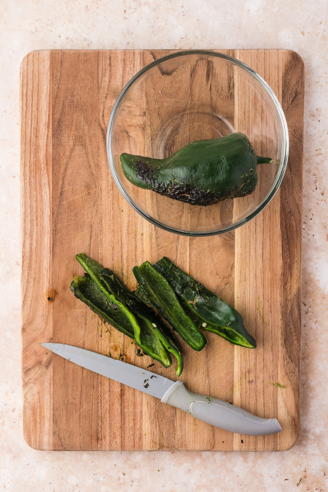 One sliced charred poblano pepper on a wooden cutting board and another whole one in a glass bowl.