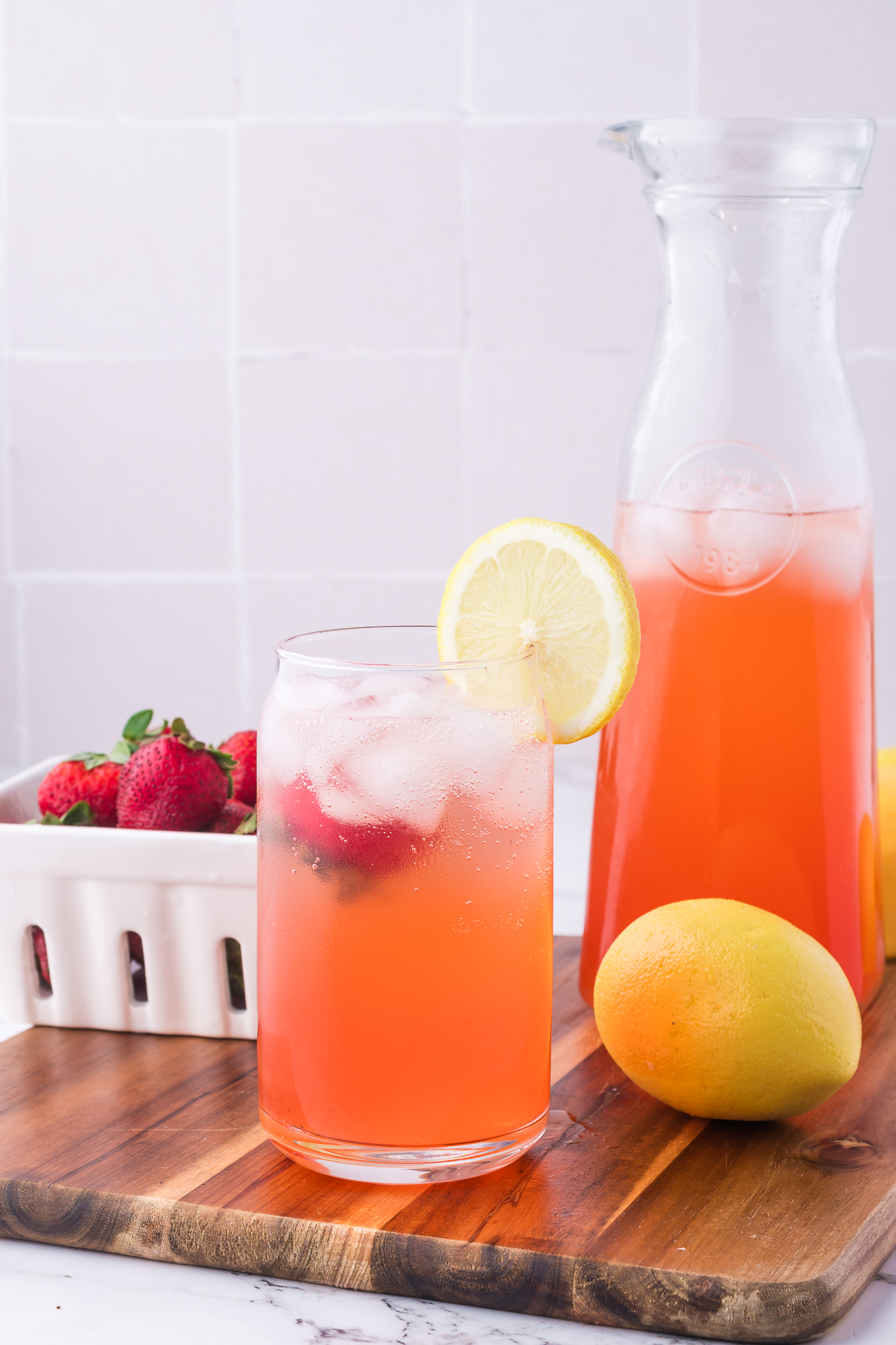 A glass of Sparkling Strawberry Lemonade with a whole strawberry and a slice of lemon on the rim next to a carafe.