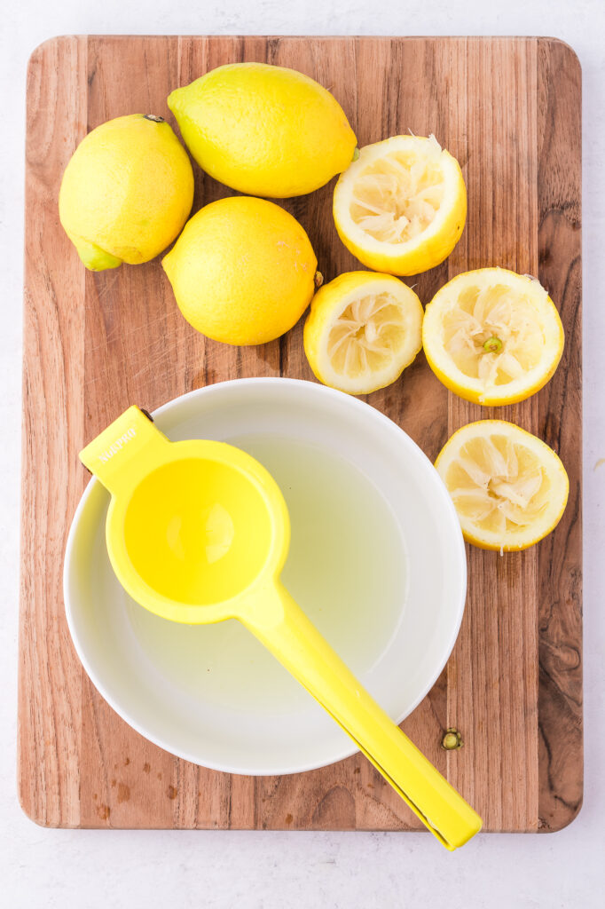 A citrus squeezer on a white bowl of fresh squeezed lemon juice, surrounded by squeezed lemons.