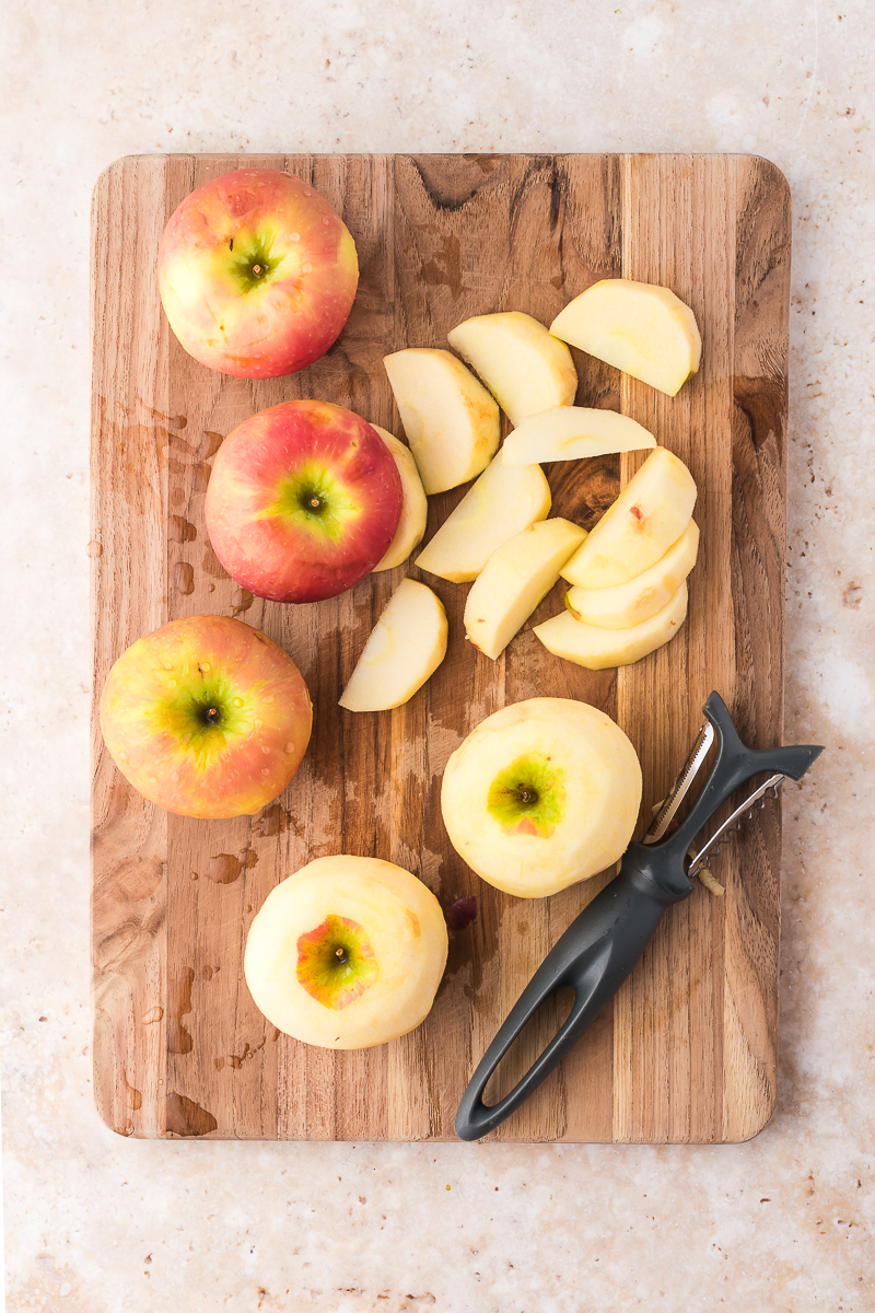 Five apples on a wooden cutting board and two of them have been peeled.