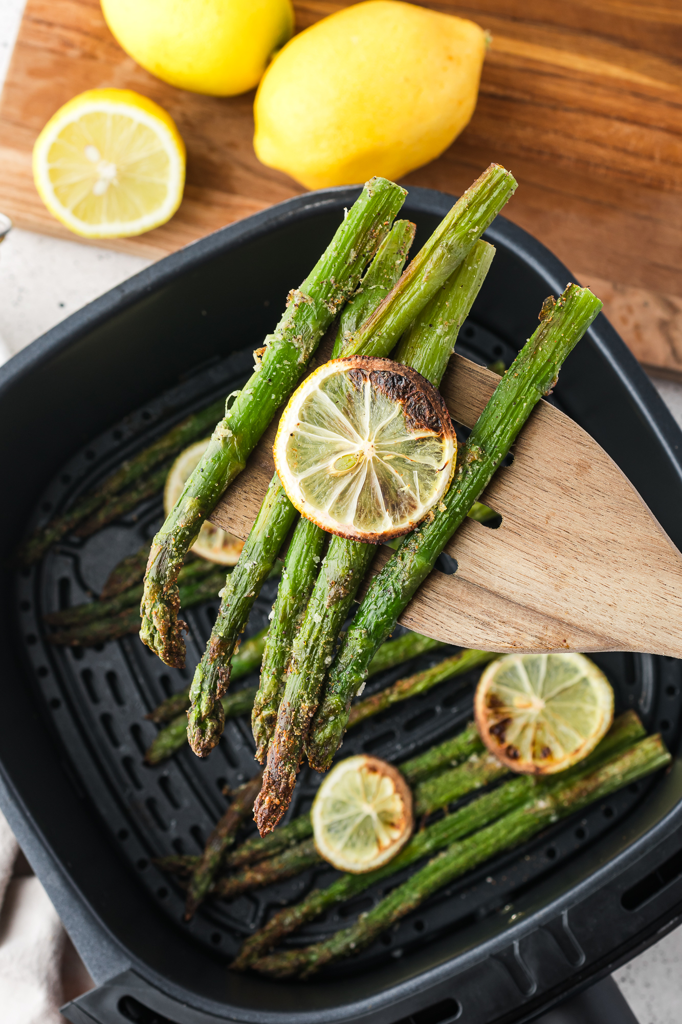 A wooden fork holding up 5 spears and one lemon slices from the air fryer basket of Air Fryer Lemon Asparagus.