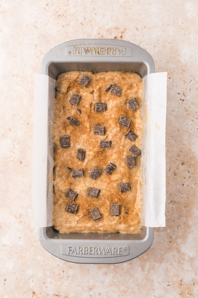 A loaf pan filled with batter for Chocolate Chunk Banana Bread topped with chocolate chunks.