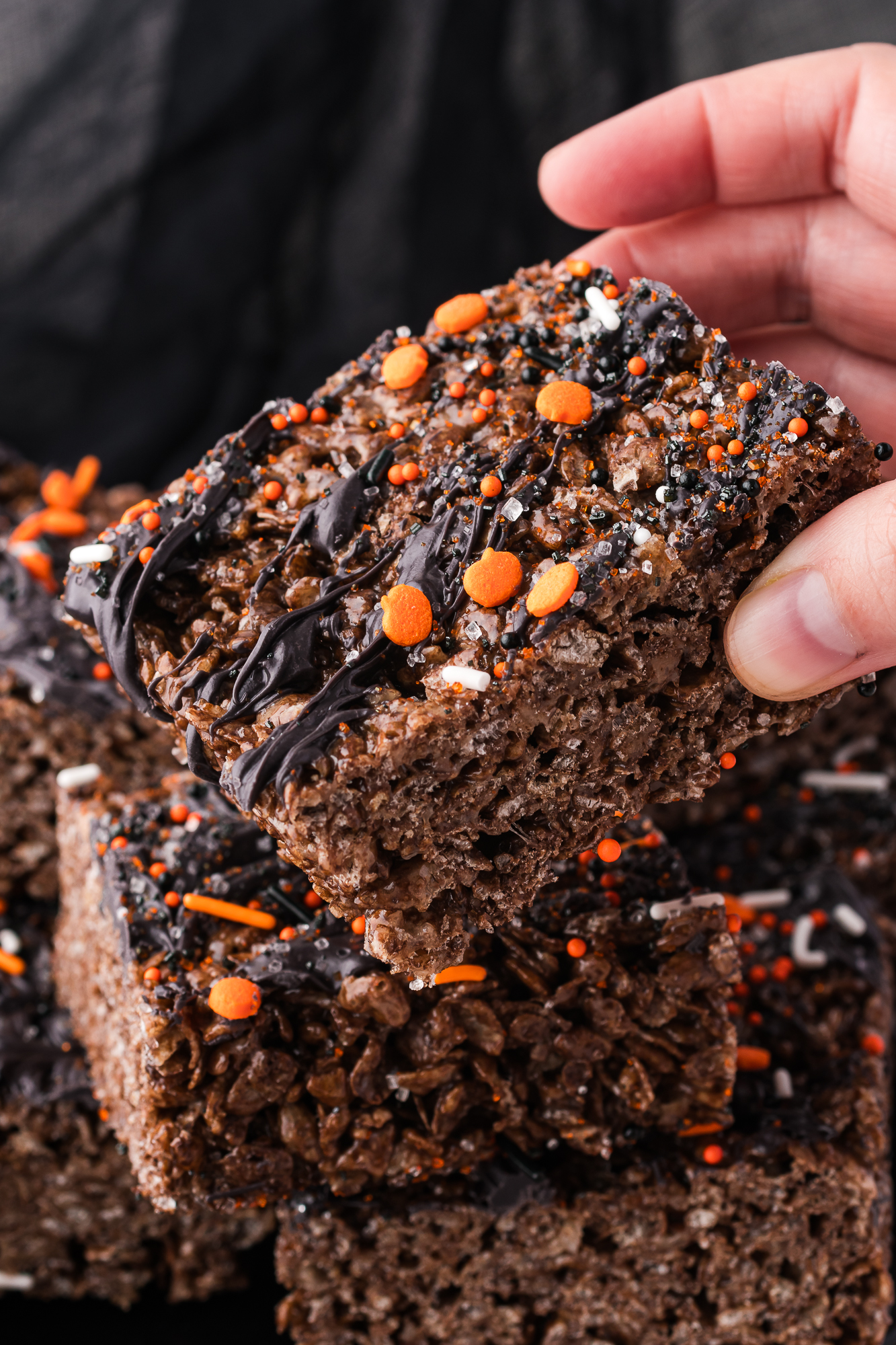 A hand picking up one piece from a pile of Spooky Cocoa Pebbles Treats.