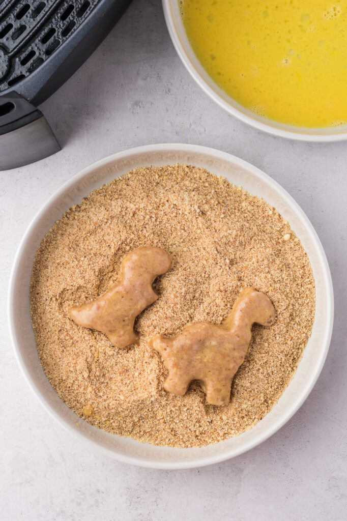 Two raw Air Fryer Dino Chicken Nuggets in the bowl of bread crumbs.