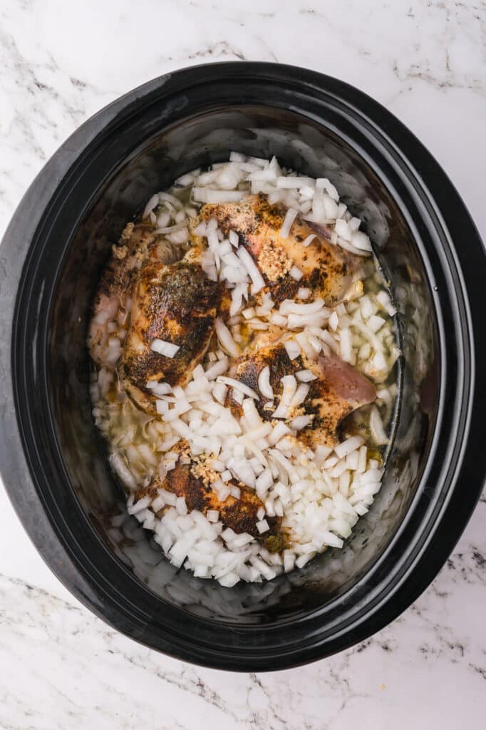 Four seared chicken breasts and diced onions in a slow cooker.