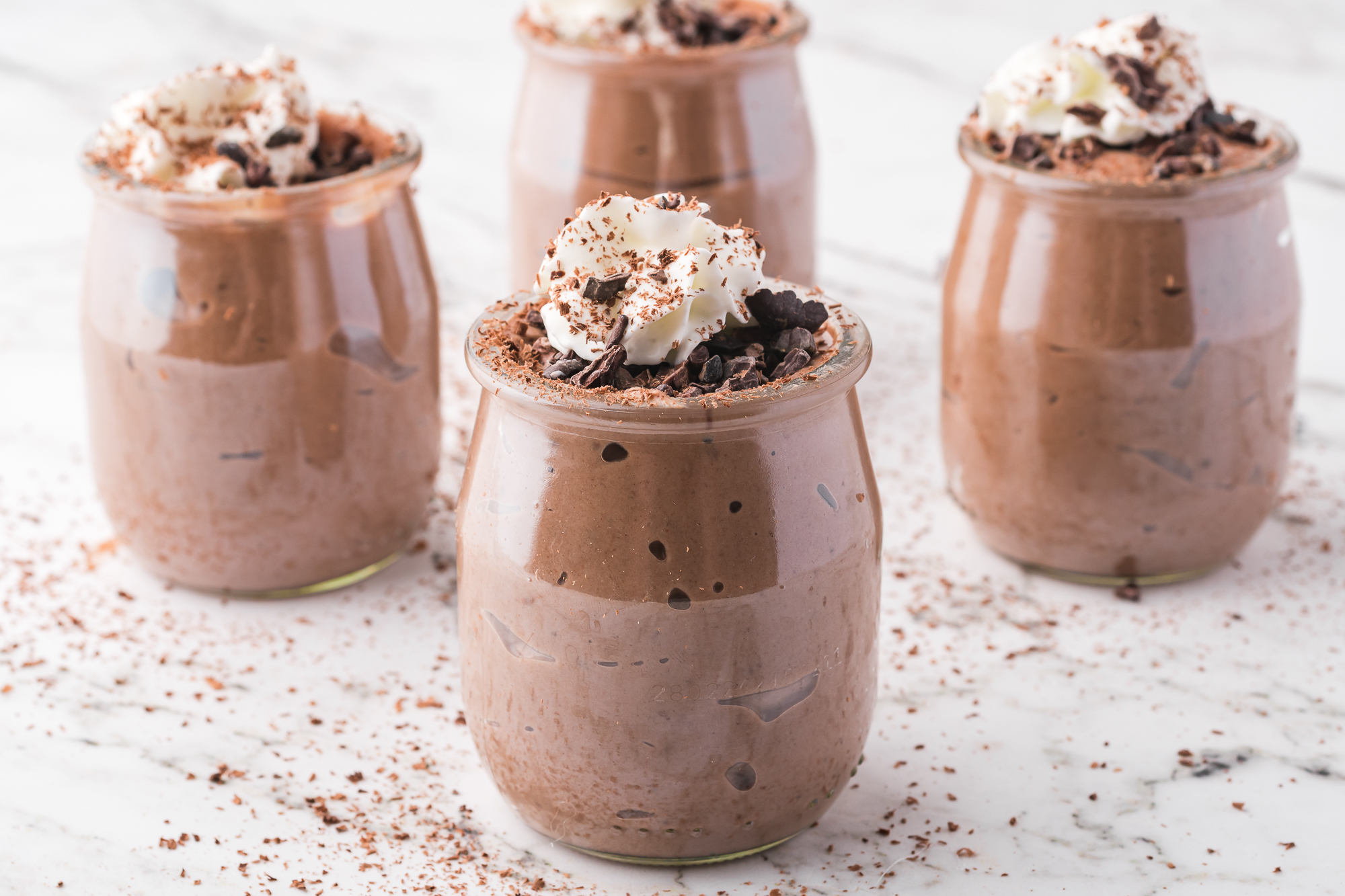 Four glasses of Chocolate Protein Pudding topped with shaved chocolate and whipped cream.