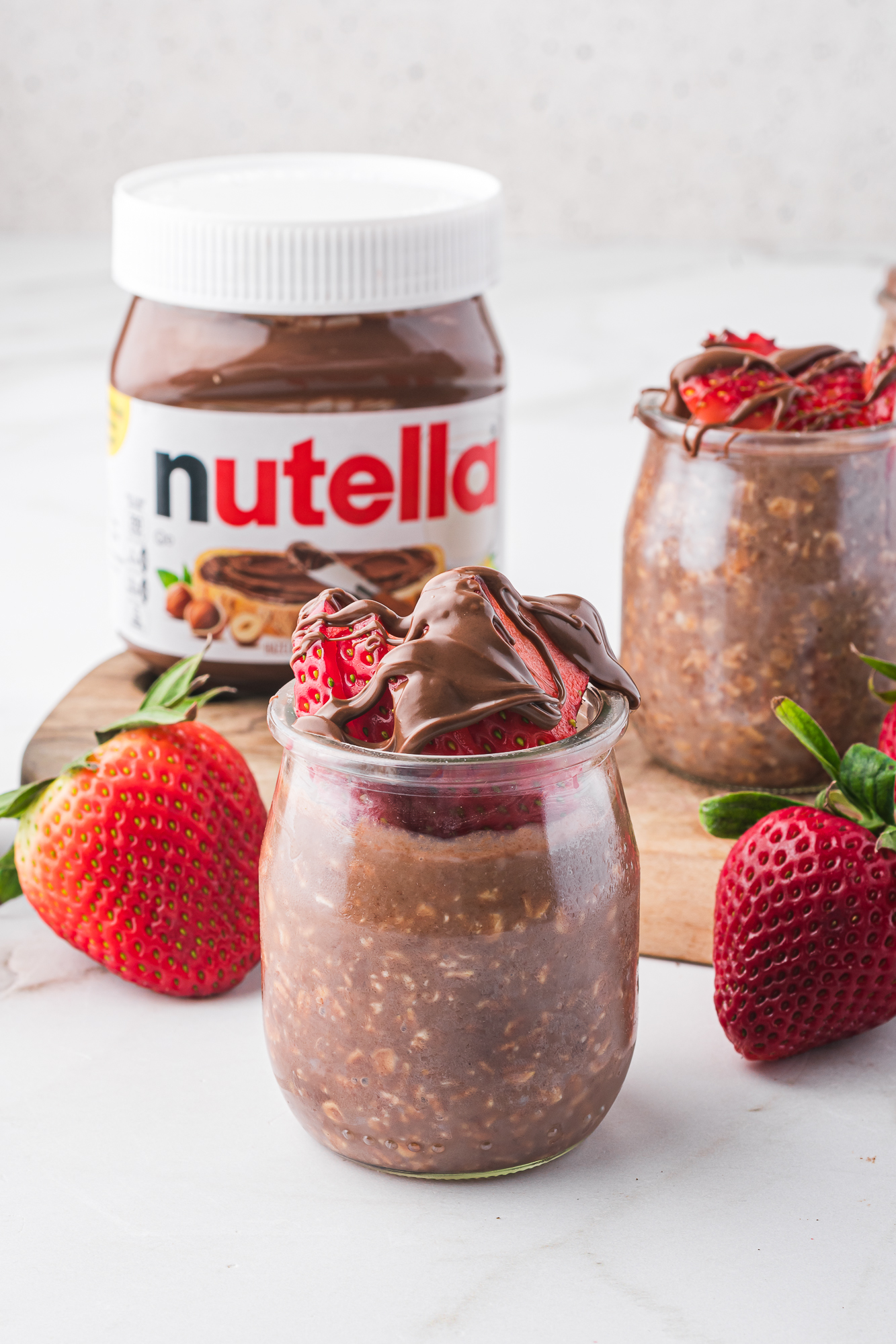 A jar of Nutella Overnight Oats topped with sliced strawberries and drizzled with Nutella standing in front of a jar of Nutella and surrounded by strawberries.