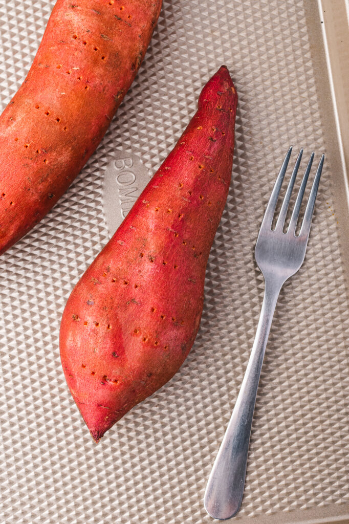 Two raw sweet potatoes on a baking sheets next to a fork.