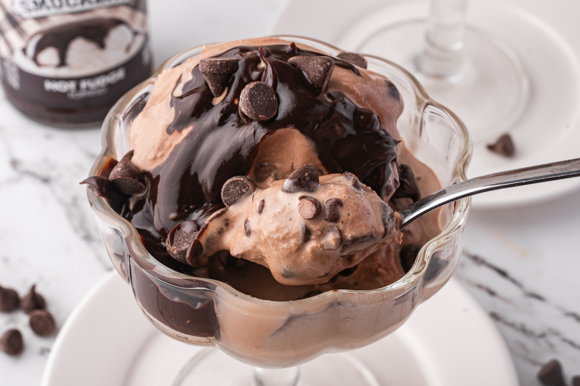 A close-up of a spoon pick up a spoonful of A glass bowl of Ninja Creami Chocolate Fudge Chip Ice Cream with fudge and chocolate chips on top.