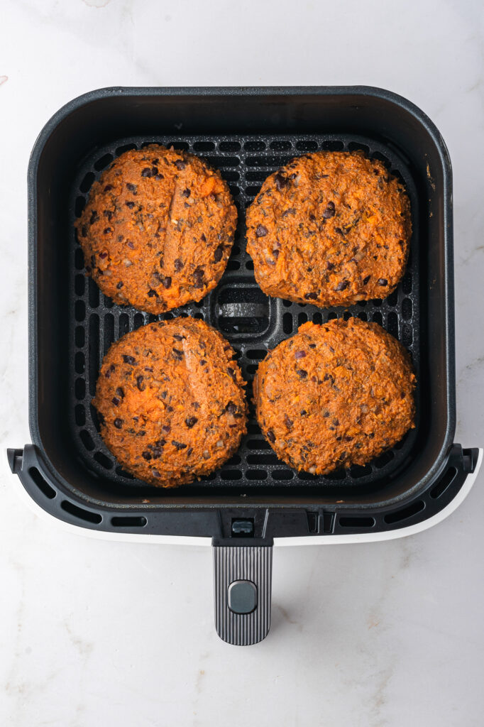 Four raw shaped patties of sweet potato and black bean mixture in the basket of an air fryer