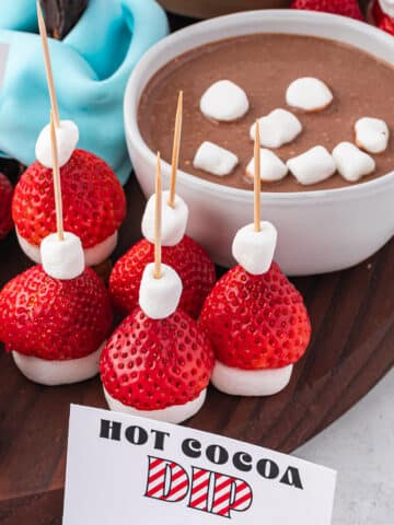 Hot Cocoa Dip in a mug with a mini marshmallow smiley face next to some santa hat strawberry skewers for dipping