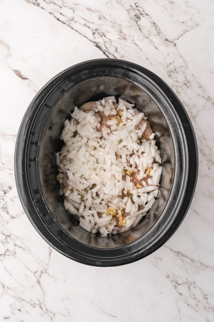A slow cooker base with raw, seasoned chicken breasts and aromatics