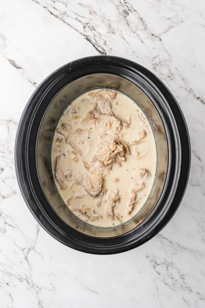 A slow cooker base with the mixed ingredients for Alfredo sauce and the shredded chicken