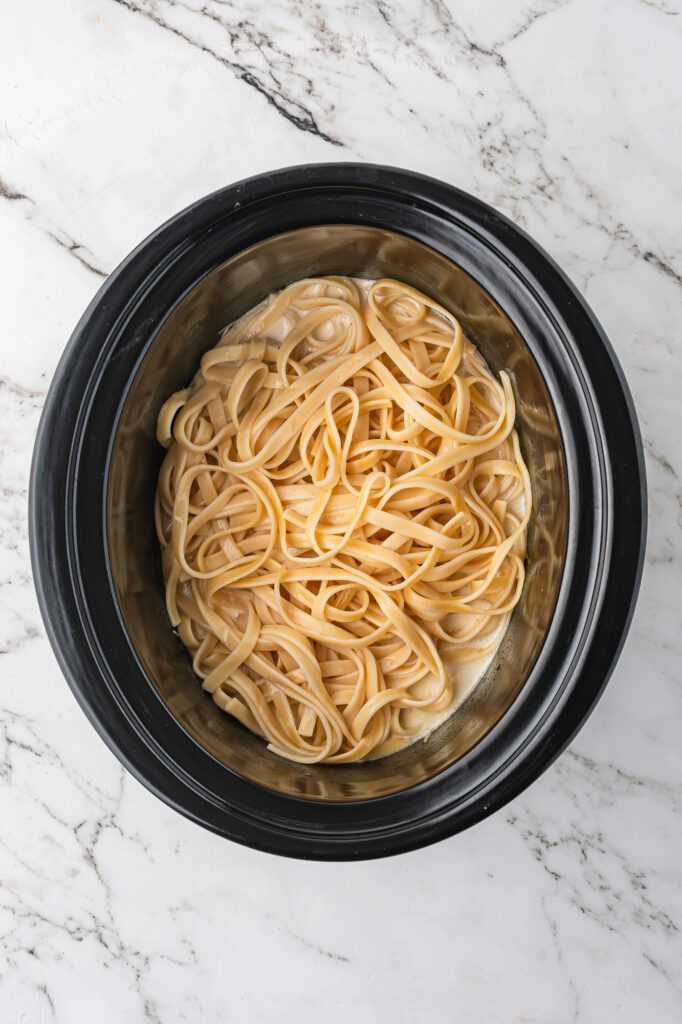 A slow cooker base with the mixed ingredients for Alfredo sauce, the shredded chicken, and pasta on top