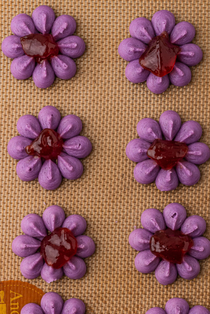 The purple wreath-shaped cookie dough shapes with a dollop of jam in the center on a silicone baking mat.