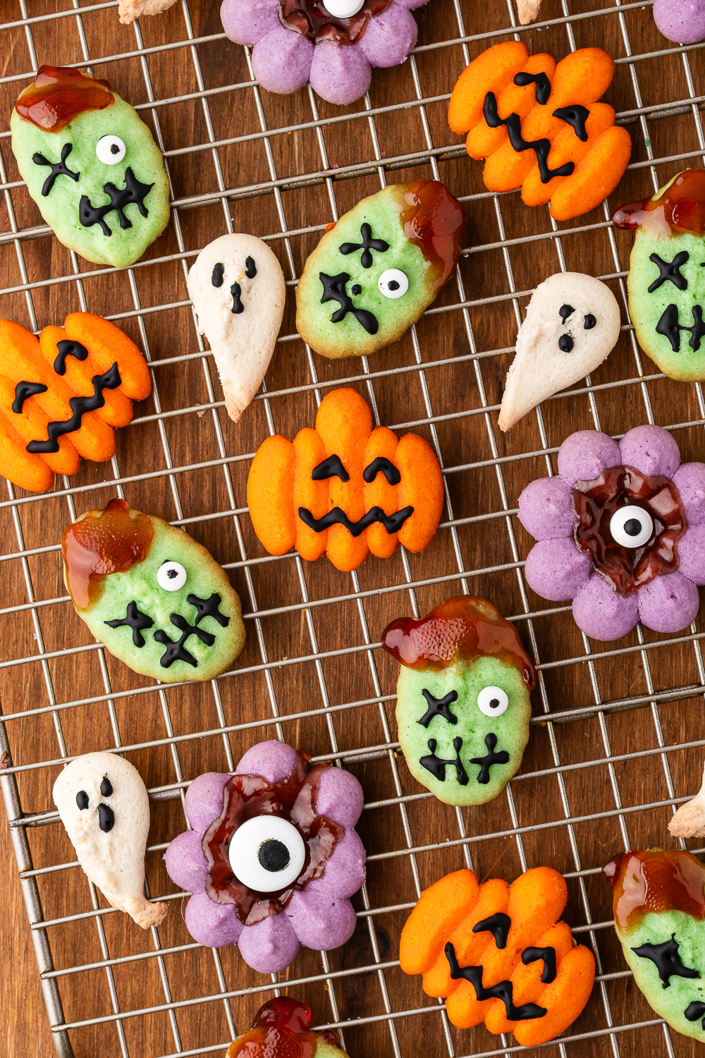A cooling rack of decorated Halloween Spritz Cookies - a variety of orange jack-o-lanterns, purple eyeball monsters, white ghosts, and green monster faces with one candy eyeball and one frosting "x" eye.