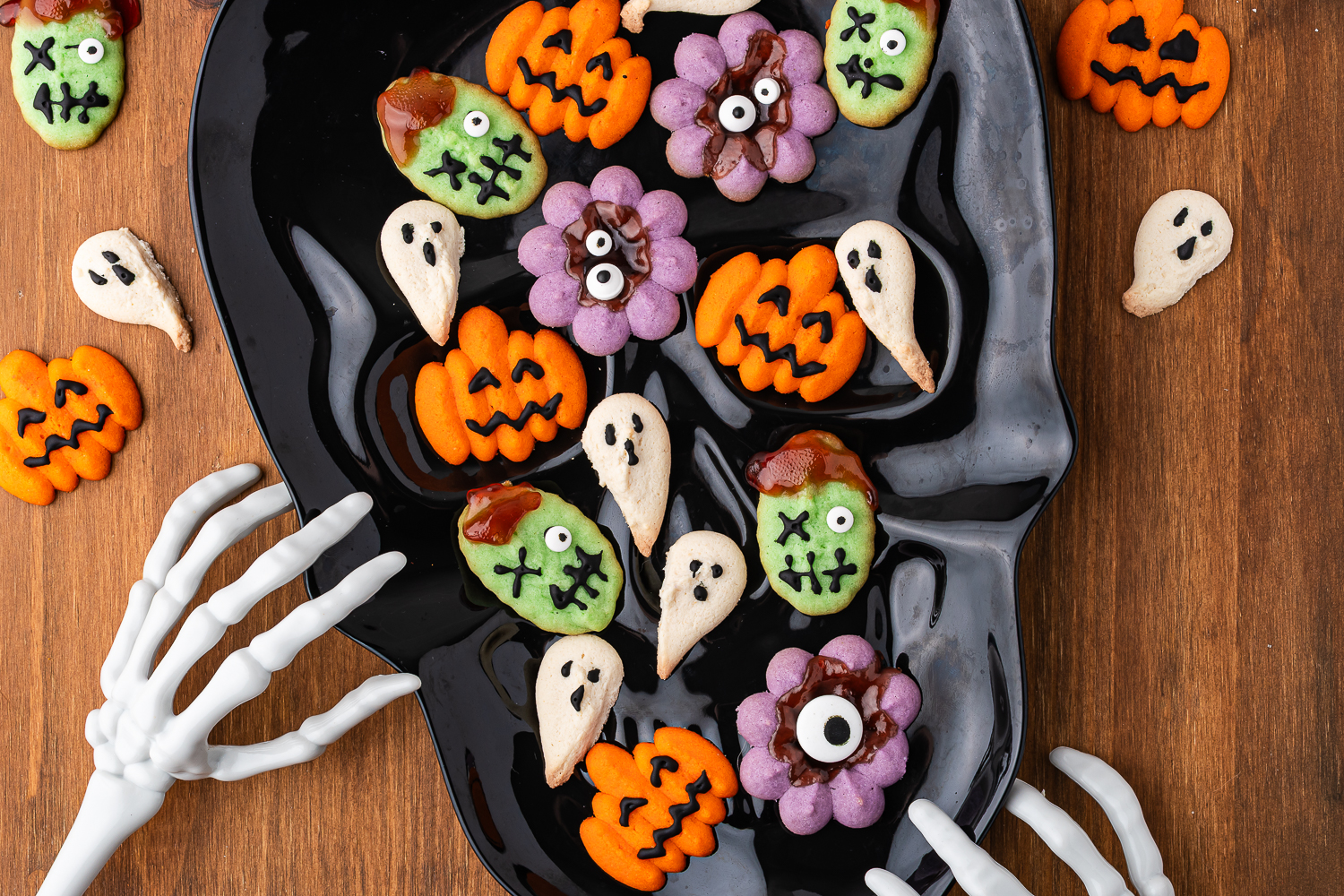 A black skull-shaped plate of decorated Halloween Spritz Cookies - a variety of orange jack-o-lanterns, purple eyeball monsters, white ghosts, and green monster faces with one candy eyeball and one frosting "x" eye. Two spooky skeleton hands decoration are reaching for the plate.