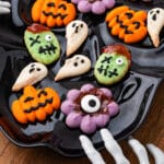 A close-up of a black plate of decorated Halloween Spritz Cookies being reached for by a spooky skeleton hand decoration.