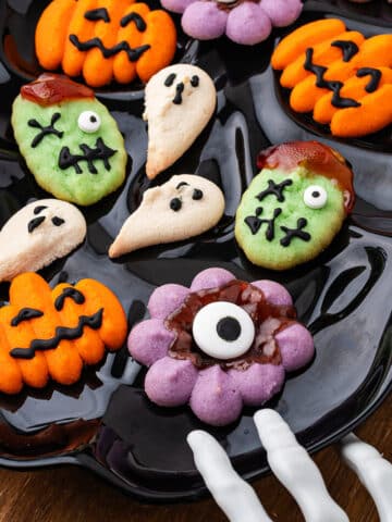 A close-up of a black plate of decorated Halloween Spritz Cookies being reached for by a spooky skeleton hand decoration.
